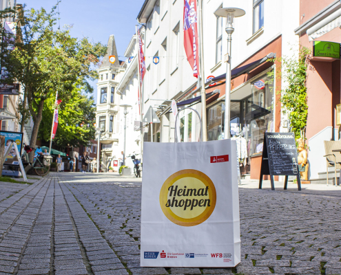 Weiße Papiertüte mit dem Logo der Aktion "Heimat shoppen". Sie steht auf dem Boden der Fußgängerzone der Vegesacker Altstadt. im Hintergund sieht man Geschäfte und Häuser